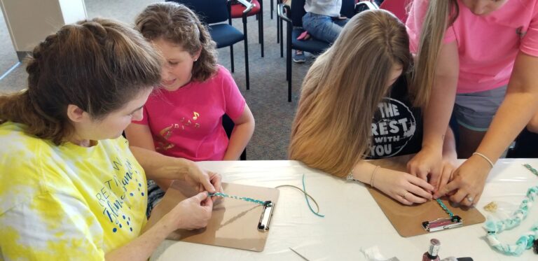 A volunteer helps two artists bead on clipboards side by side, nail polish and beading tools on the table.