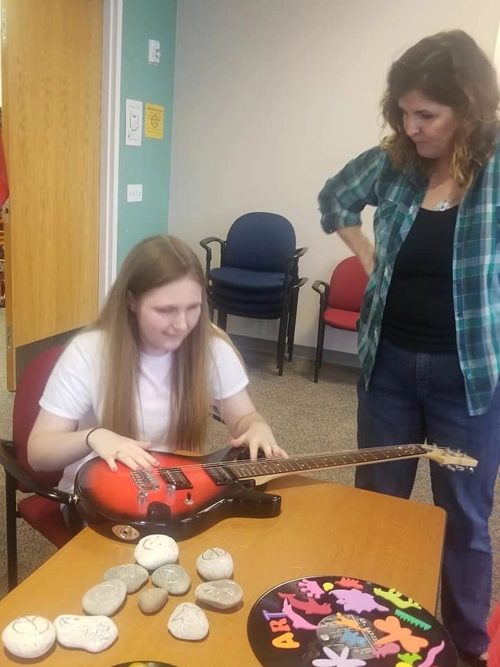 An artist runs a hand across an electric guitar decorated with painted stones displayed beside it, a volunteer looking on.