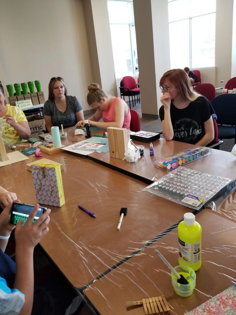 Artists sit around a table decorating wooden boxes with paint and gems, rhinestone sheets and a card reading Happy Mother's Day nearby.