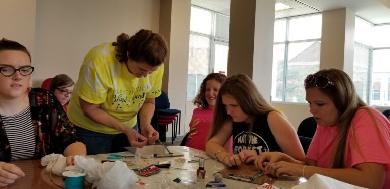 A volunteer helps a group of artists bead at a table covered in nail polish, small trays and craft supplies.