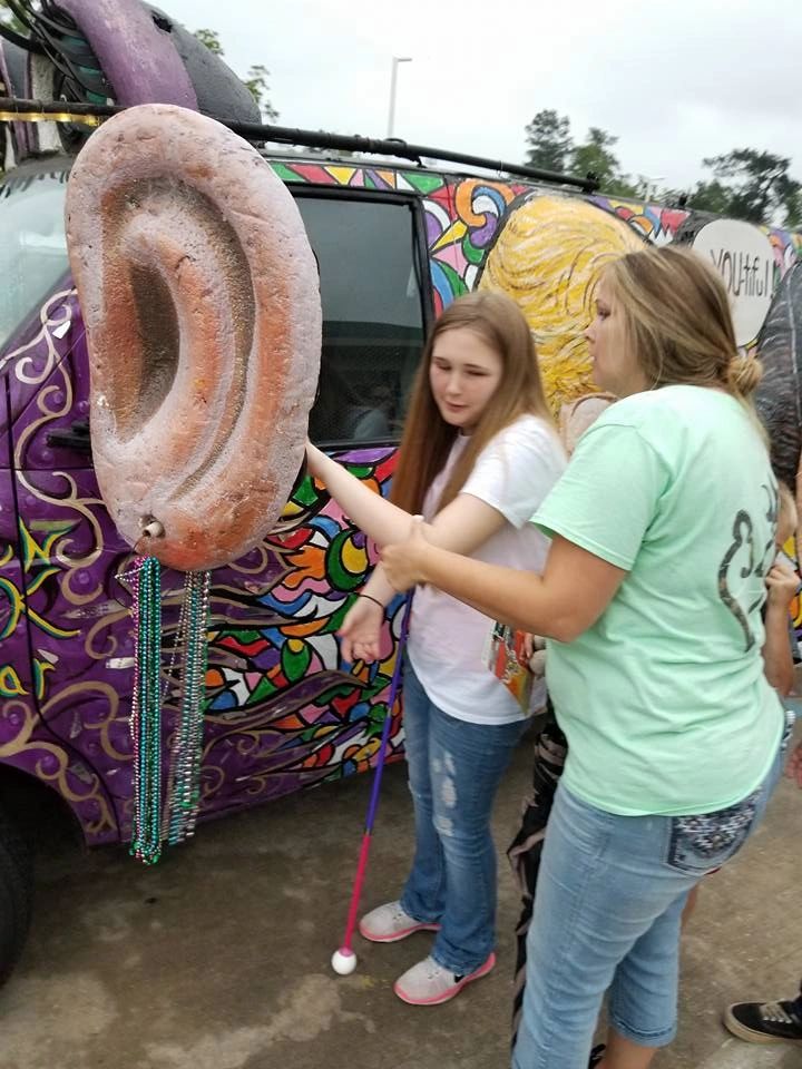 A volunteer guides an artist's hand to touch the large sculpted ear on the Women Rock Art Car, with others gathered around.