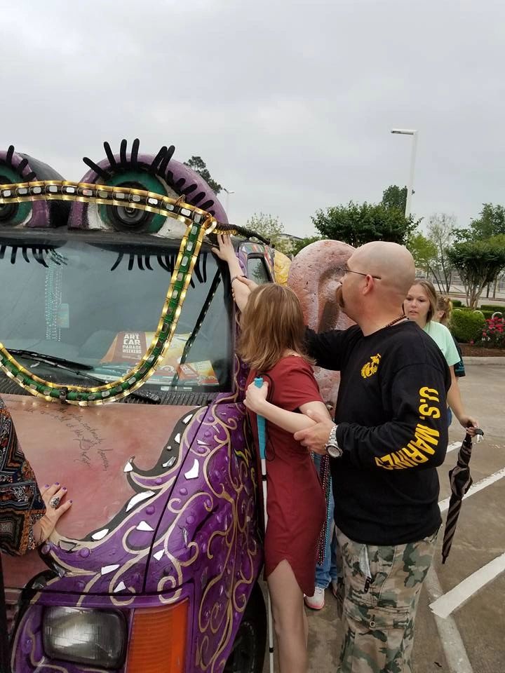 A volunteer holds an artist close while they reach up to touch the sculpted ear on the front of the Women Rock Art Car, a painted nose visible beside them.