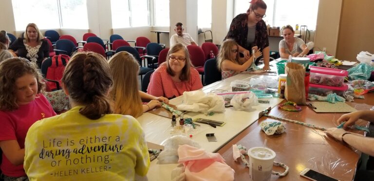 Two artists laugh together at a table while beading, one wearing a pink shirt, a volunteer in yellow helping across from them.
