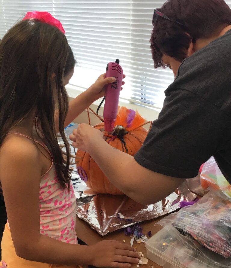 An artist uses a heat tool to dry a decorated pumpkin trimmed with orange feathers and a small bell.