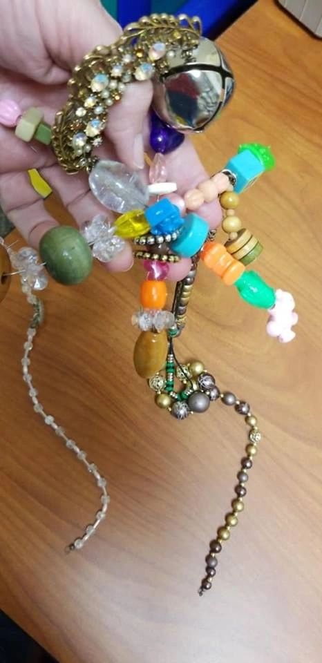 A hand holds a cluster of beaded charms and pendants strung together, including a large silver bell, glass beads and small figures.