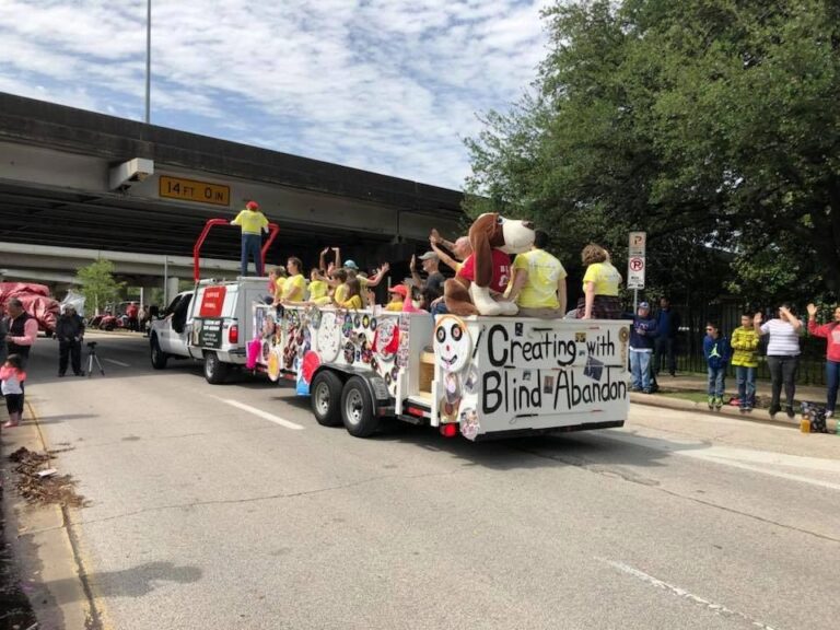 The float travels beneath an overpass, with a giant stuffed hound in a Blind to the Bone shirt riding at the front and Creating With Blind Abandon painted across the panel.