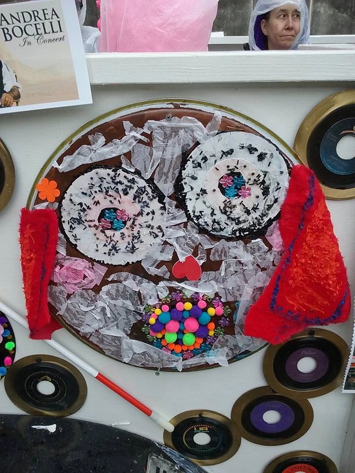 A decorated record made to look like a face, with sprinkle-textured circles for eyes, red felt eyebrows and a cluster of pom-poms for a mouth, mounted on the float alongside more records and a white cane.