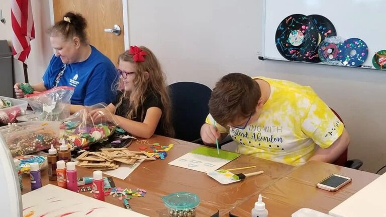 Artists sit at a table decorating wooden picture frames and painting, with more decorated records displayed on a whiteboard behind them.