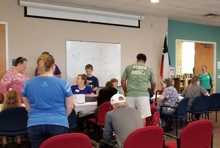 A wide view of the studio, with artists and volunteers gathered around several tables, a whiteboard sketched with a tree design and a Texas flag near the doorway.