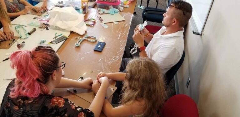 A volunteer helps two artists at a table, one working with beaded cord, colourful materials spread across the tabletop.