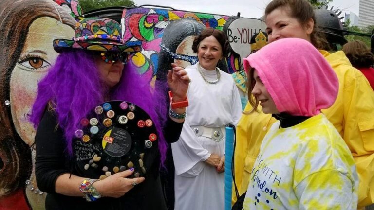 A person in a purple wig and hand-painted hat holds a beaded braille tag near a woman dressed as Princess Leia, both smiling in front of the parade car's colourful painted panels.