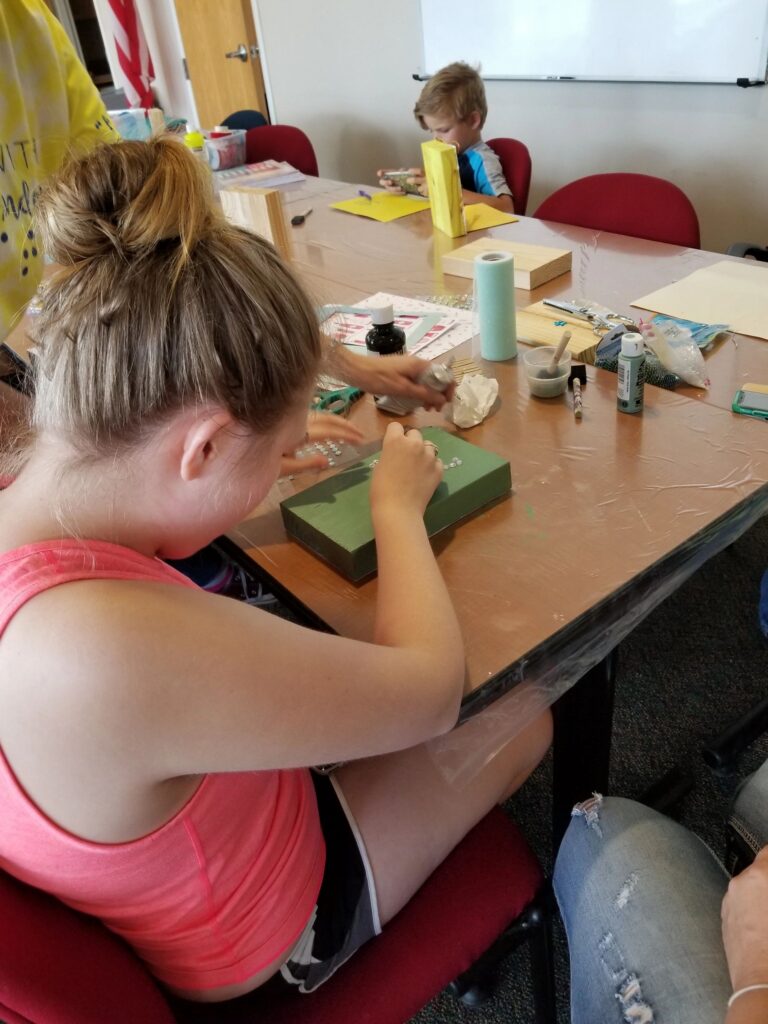 Artists sit around a table making beaded bracelets, one wearing a shirt printed with a Helen Keller quote, craft bags and beads spread across the surface.