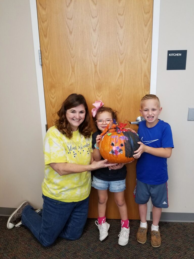 A volunteer and two artists hold up a pumpkin decorated with a painted face, gem accents and colourful feather trim.