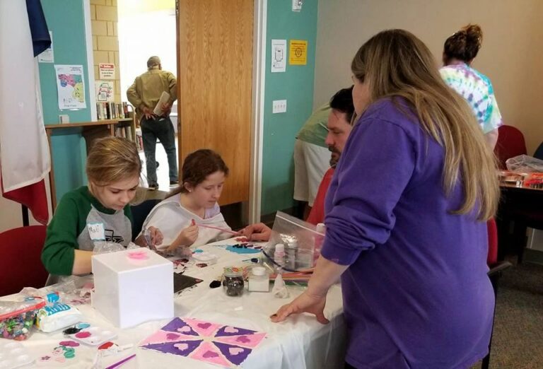 A volunteer helps two artists at a table set with glitter, small boxes and paper cut into heart shapes.