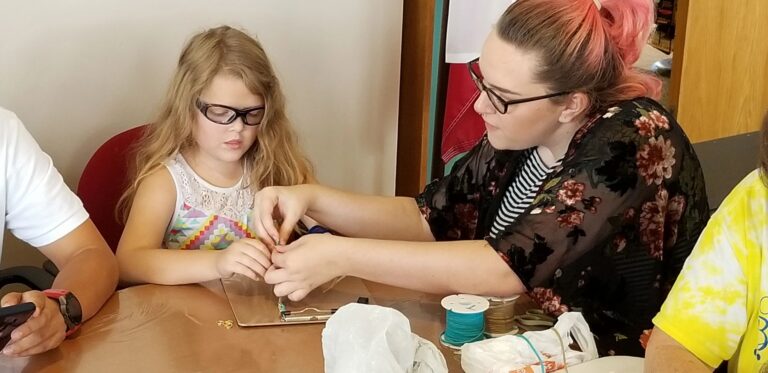 A volunteer helps a young artist bead on a small board, thread spools and craft supplies nearby on the table.