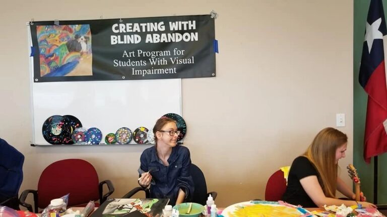 An artist chats at a table beneath a banner reading Creating With Blind Abandon, with decorated records displayed on a whiteboard behind them.