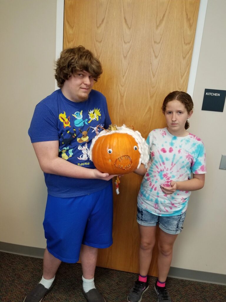 Two artists hold up a pumpkin wrapped in white yarn with googly eyes and gold-studded accents.