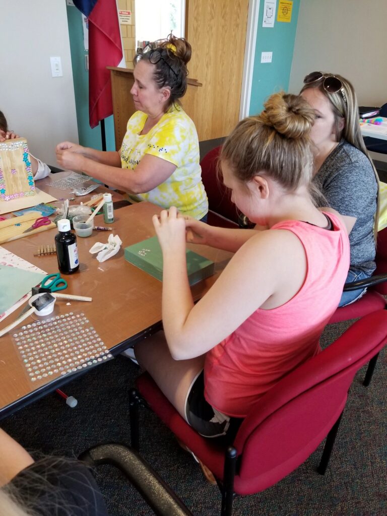 Artists sit at a table decorating wooden pieces with gems and glue, a whiteboard visible in the background.