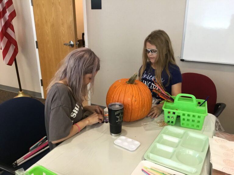 An artist paints a whole pumpkin at a table set with paint trays and a green supply basket.
