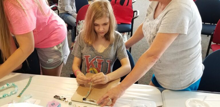 A volunteer helps an artist bead on a clipboard, more beading materials and small trays visible on the table.