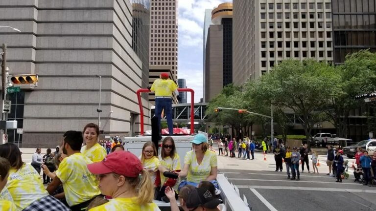 Riders wave from the top platform of the truck towing the float as it moves through downtown, tall buildings lining the street.