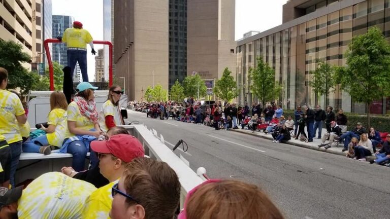 Riders sit atop the float as it passes through downtown, the towing truck's raised platform visible ahead against the skyline.