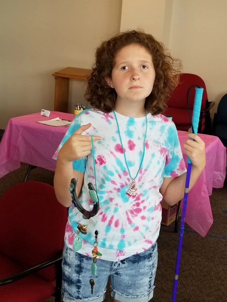 An artist stands holding a beaded necklace with a horseshoe pendant, a white cane in the other hand.
