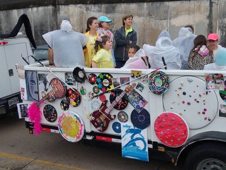 A rider on the float holds a white cane braced against the railing, with decorated records and a truck marked Service Animal visible alongside.