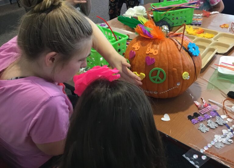 Two artists decorate an orange pumpkin with a peace sign, foam flowers and small gems.