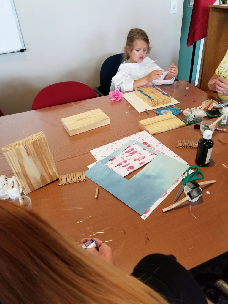An artist works on a wood block decorated with small flower gems, a pink fabric rose resting nearby on the table.