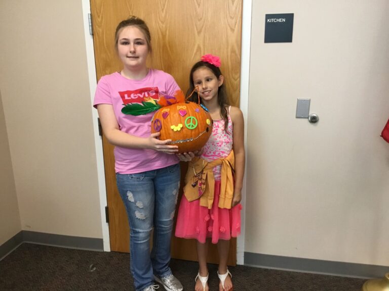 Two artists hold up a pumpkin decorated with a peace sign, painted flowers and gem accents.