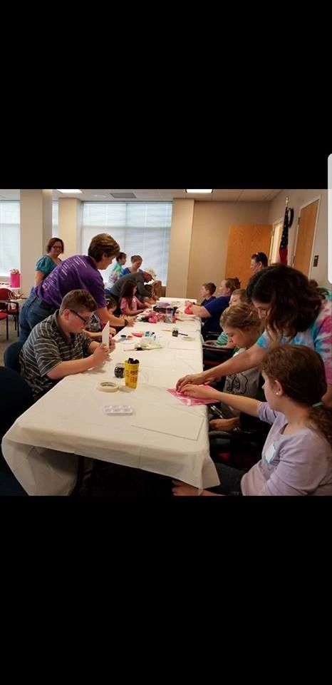 A wide view of the studio, with artists and volunteers seated around a long table working on a craft project with paint palettes and tape.