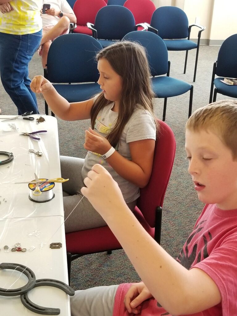 Two artists thread beads onto wire together at a table with horseshoes and charms nearby.