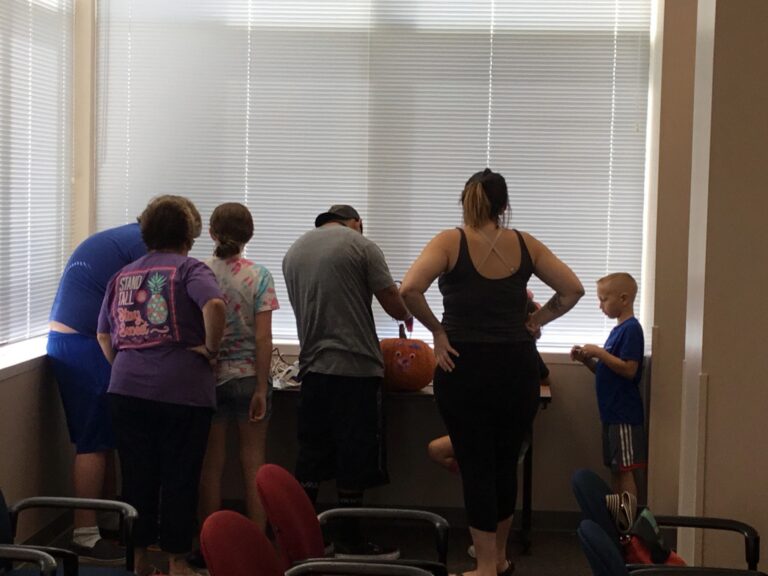A group stands at a window ledge, one placing a small decorated pumpkin on the sill.