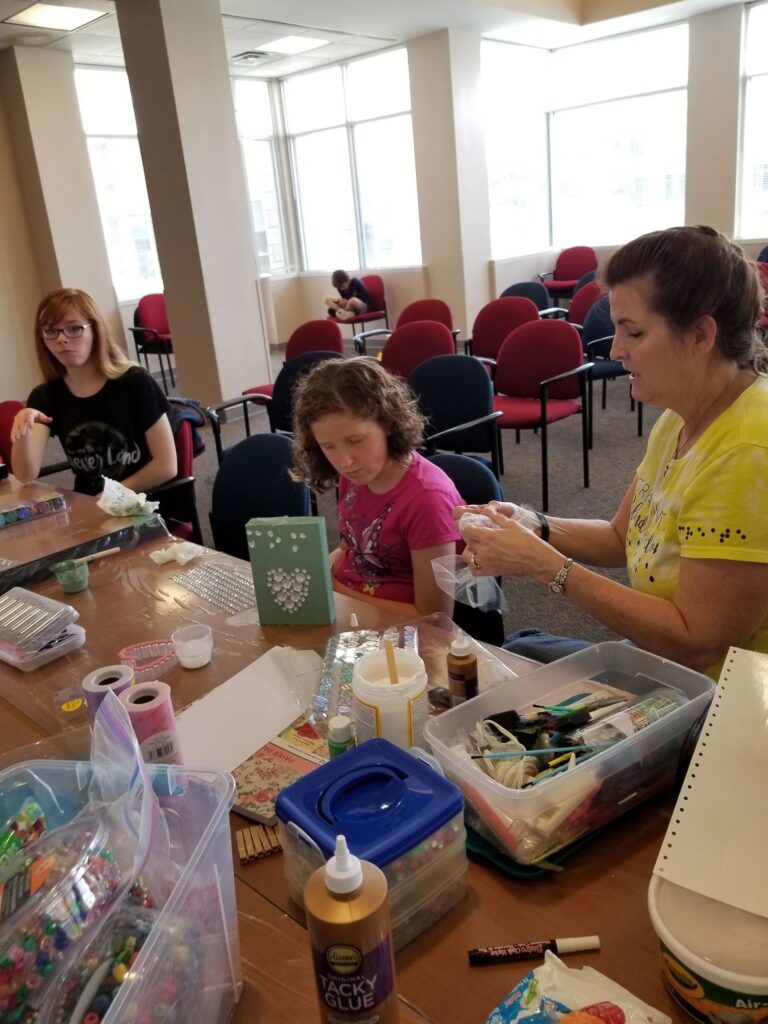 Artists sit at a table decorating a mint green box with pearl-drop rhinestones, craft trays and glue nearby.