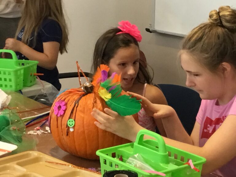 Two artists work together attaching a green feather to a pumpkin decorated with a foam flower and smiley sticker.