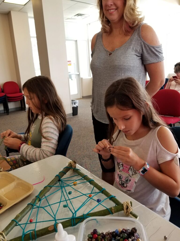 A volunteer helps two artists weave beads onto a triangular stick-frame dream catcher at a table covered in beads.