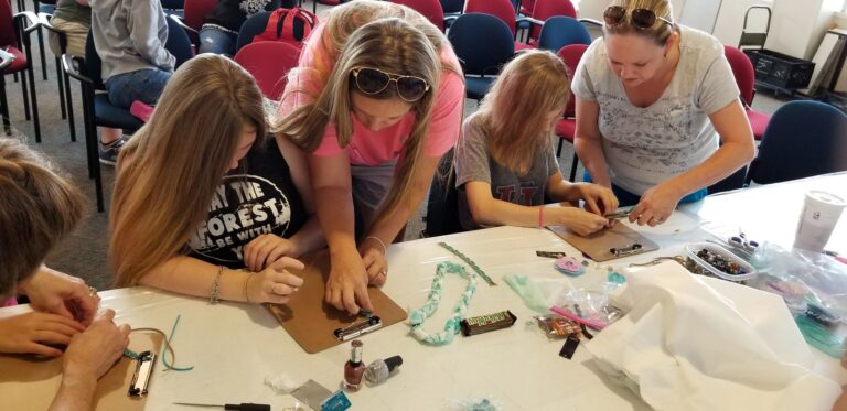 Several artists and a volunteer bead together at a table covered in cord, nail polish and small trays of supplies.