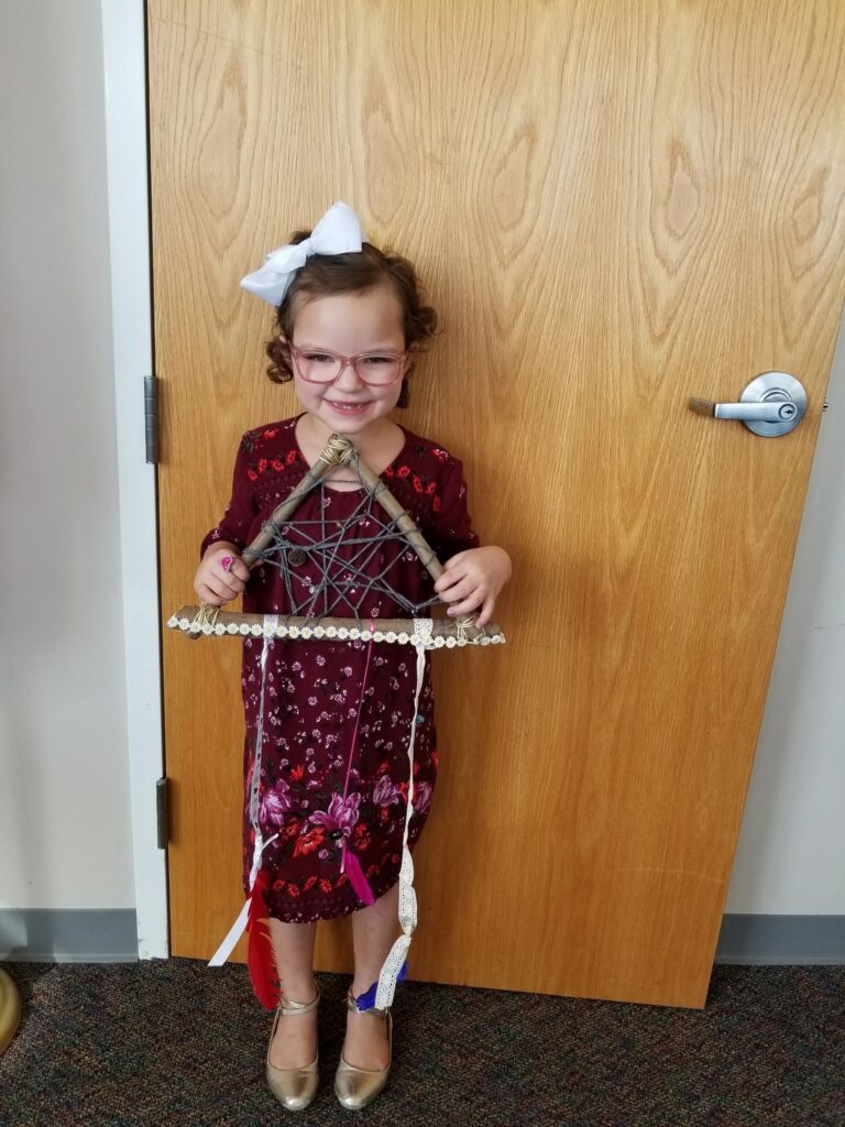 An artist holds up a finished dream catcher woven with dark yarn between three wooden dowels, trimmed with lace and hanging ribbons.