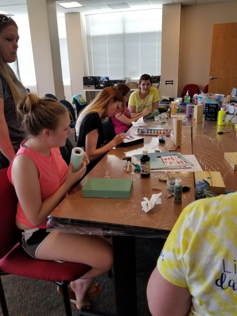 An artist decorates a green box with rhinestones at a table set with paint, ribbon and craft supplies.