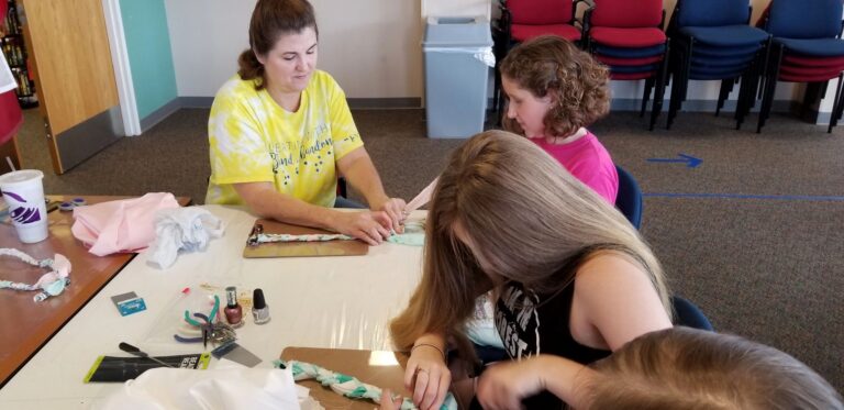 Artists and volunteers braid strips of pastel fabric into ropes at a table set with nail polish and craft supplies.