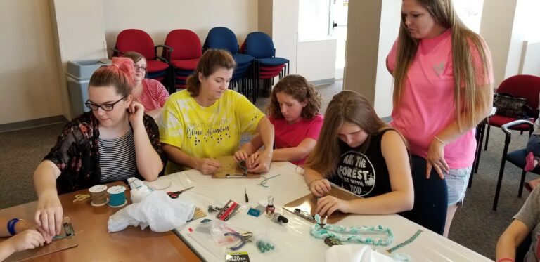 An artist twists beads onto a wire at a table, a volunteer and another artist working close by with braided fabric ropes nearby.