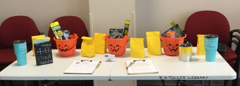 A table set up with pumpkin-shaped treat buckets, yellow gift bags and small prize items, name cards laid out beside them.