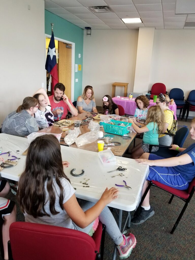 A large group of artists and volunteers sit around two tables decorating horseshoes with beads, more supplies spread across a second table.