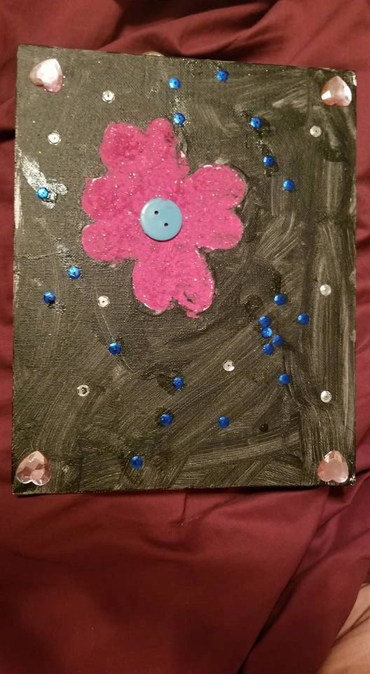 A canvas dabbed with orange, blue and green paint spots, resting on a table beside a red glitter heart sticker and a set of paintbrushes.
