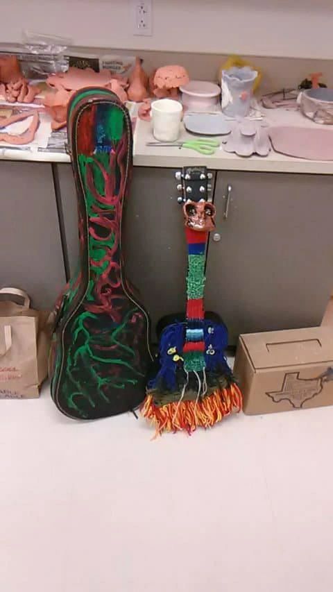 A hand-painted guitar case with red and green root-like patterns beside a small decorated guitar wrapped in colourful yarn with a painted face on its headstock, on a classroom counter with clay sculptures.