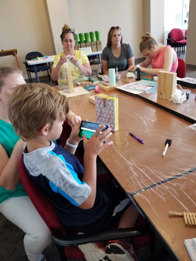 Artists sit at a table with wood blocks and craft supplies, one holding a phone, gift bags and paint bottles spread around them.
