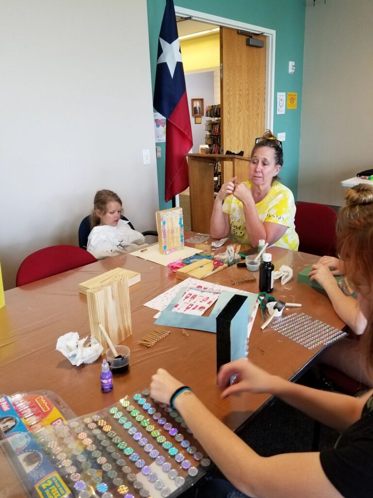 Artists sit at a table decorating wooden boxes, one holding a wood block, sheets of gems and rhinestones spread across the tabletop.