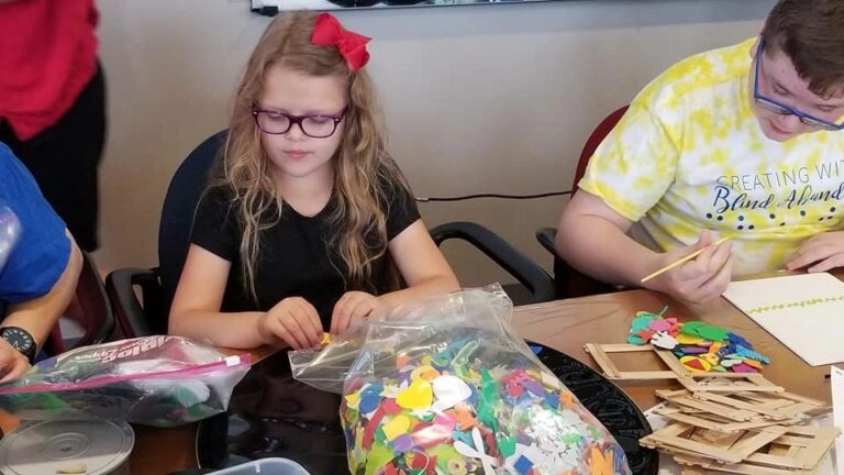 An artist sorts through a bag of colourful foam shapes at a table, while another artist paints beside them, wooden picture frame pieces spread nearby.