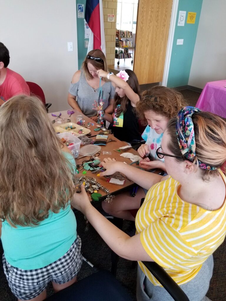 A group of artists and volunteers work together at a table covered in beads, charms and horseshoes.